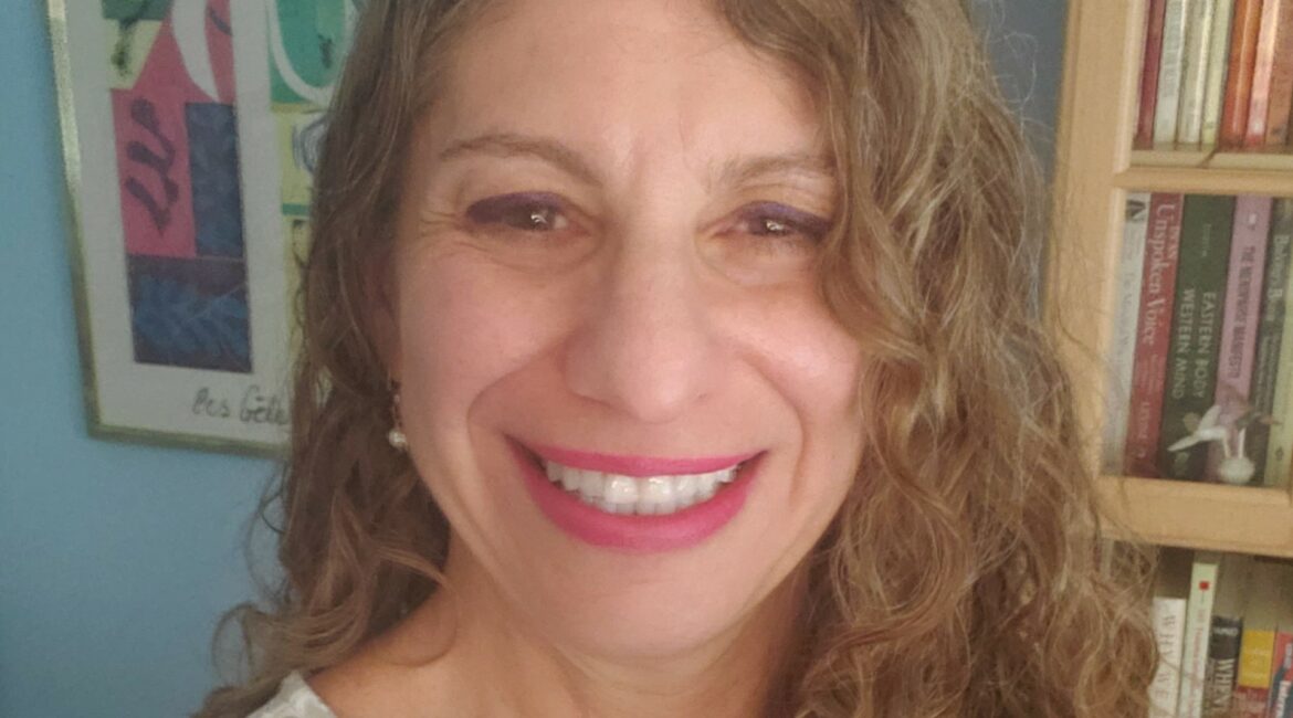 Trauma Solutions: A woman with long curly hair, wearing a white patterned top, smiles at the camera in a room with a bookshelf and abstract artwork in the background.