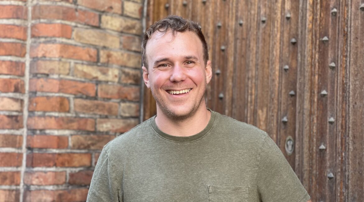 Trauma Solutions: A person with short hair wearing a green t-shirt stands smiling in front of a brick wall and a wooden door with metal studs.