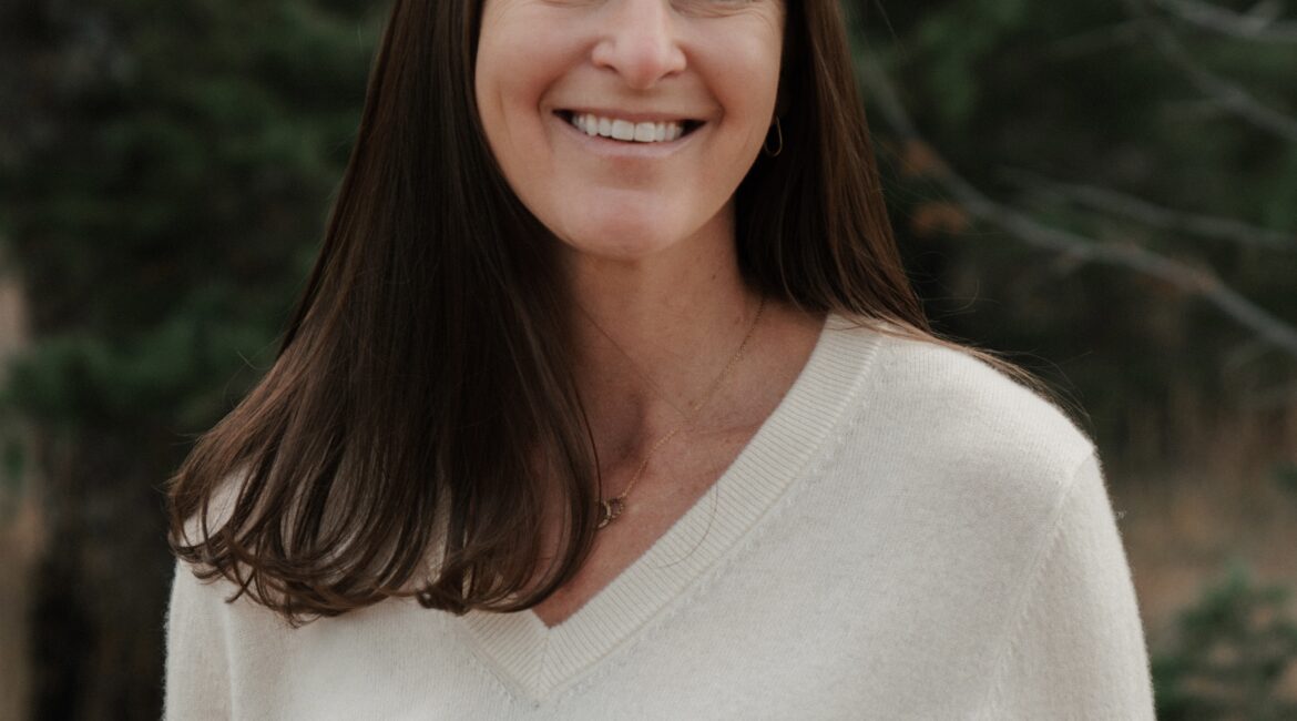 Trauma Solutions: A woman with long brown hair and a white sweater smiles outdoors with trees in the background.