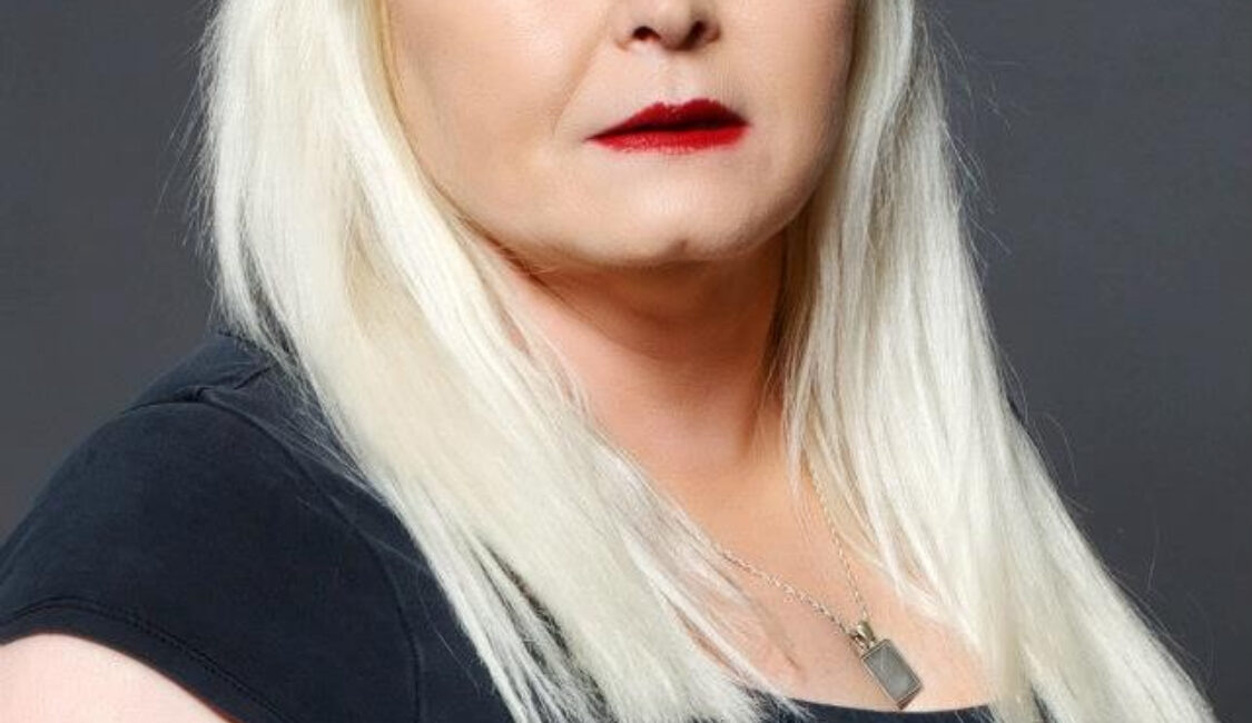 Trauma Solutions: A woman with straight, light blonde hair and bangs, wearing a black top and a necklace, poses against a plain gray background.