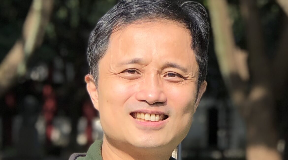 Trauma Solutions: A man wearing a striped shirt and jacket smiles outdoors with trees in the background.