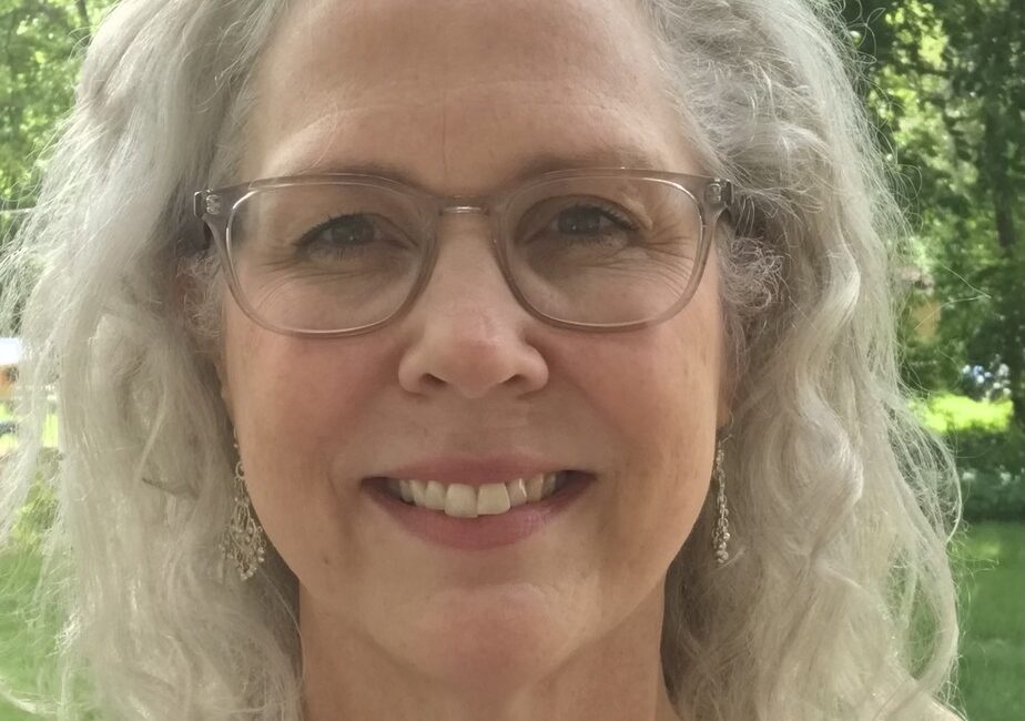 Trauma Solutions: A woman with gray curly hair, glasses, and earrings is smiling outdoors. Green trees and grass are visible in the background.