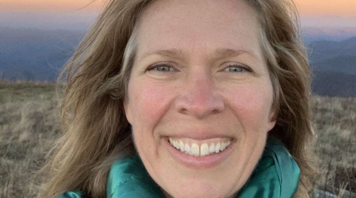 Trauma Solutions: A woman with long light hair smiles at the camera outdoors, wearing a teal jacket and backpack, with mountains and a colorful sky in the background.