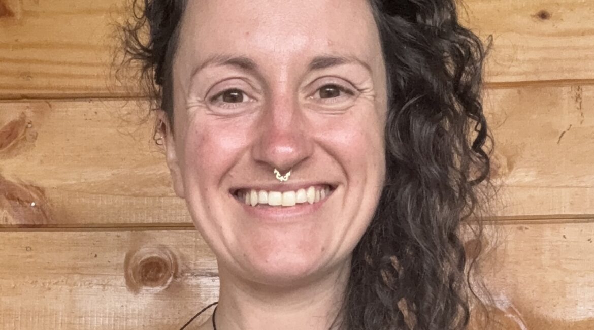 Trauma Solutions: A person with long curly hair and a nose ring smiles at the camera, standing in front of a wooden wall.
