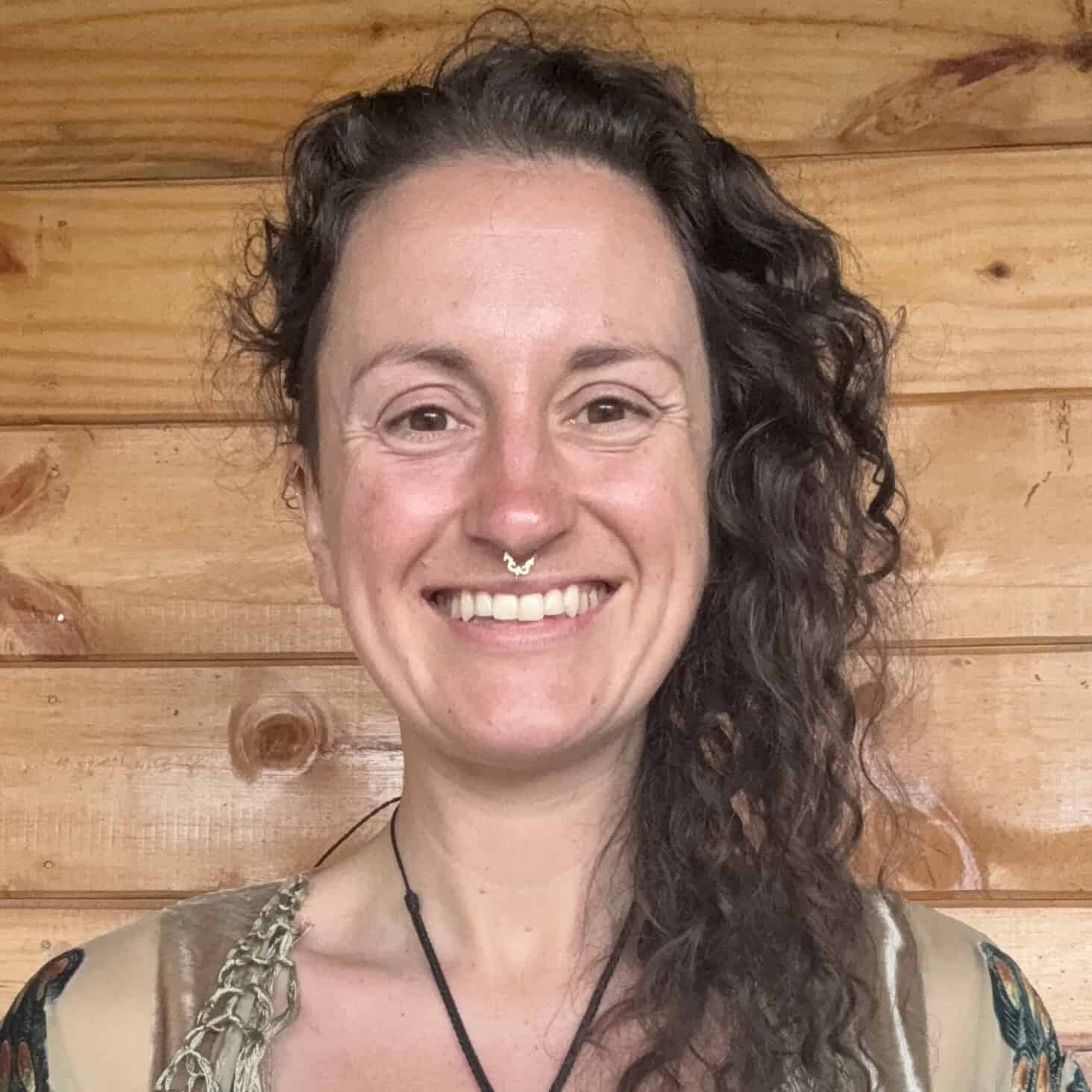 Trauma Solutions: A person with long curly hair and a nose ring smiles at the camera, standing in front of a wooden wall.