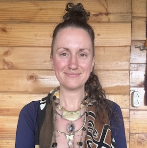 Trauma Solutions: Person with curly hair in a bun, wearing necklaces and patterned clothing, stands in front of a wooden wall with a visible electrical outlet.