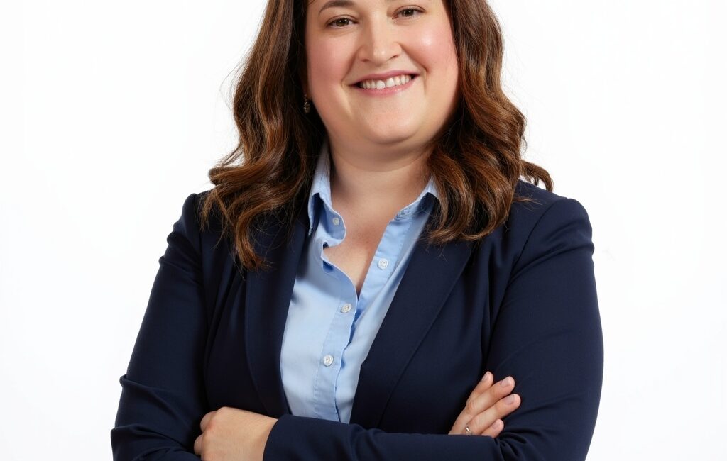 Trauma Solutions: Woman with wavy brown hair wearing a navy blazer and light blue shirt, smiling with arms crossed against a plain white background.