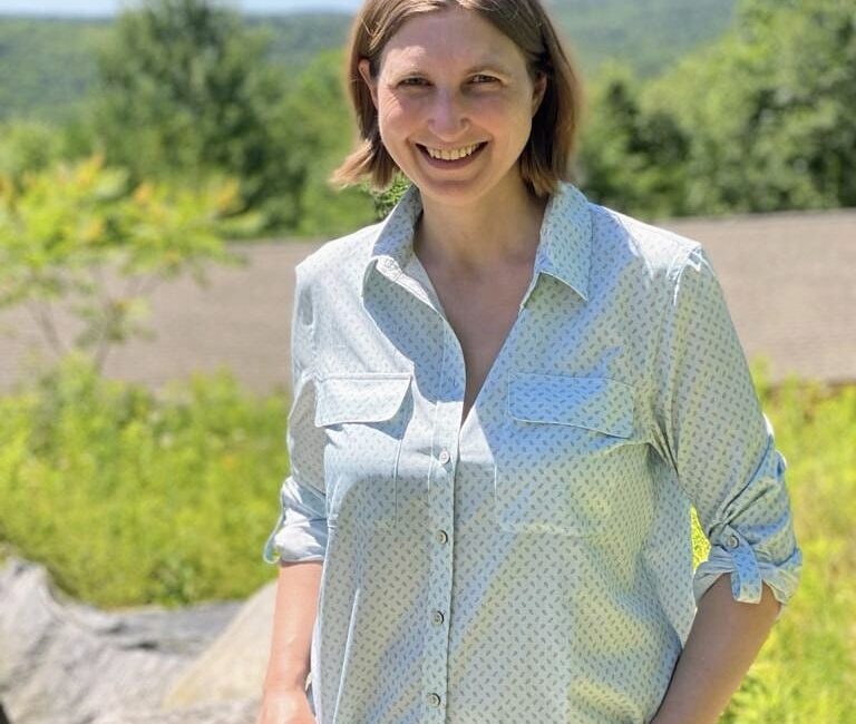 Trauma Solutions: A woman stands outdoors in sunlight, smiling, with greenery and a distant tree-covered hill in the background.