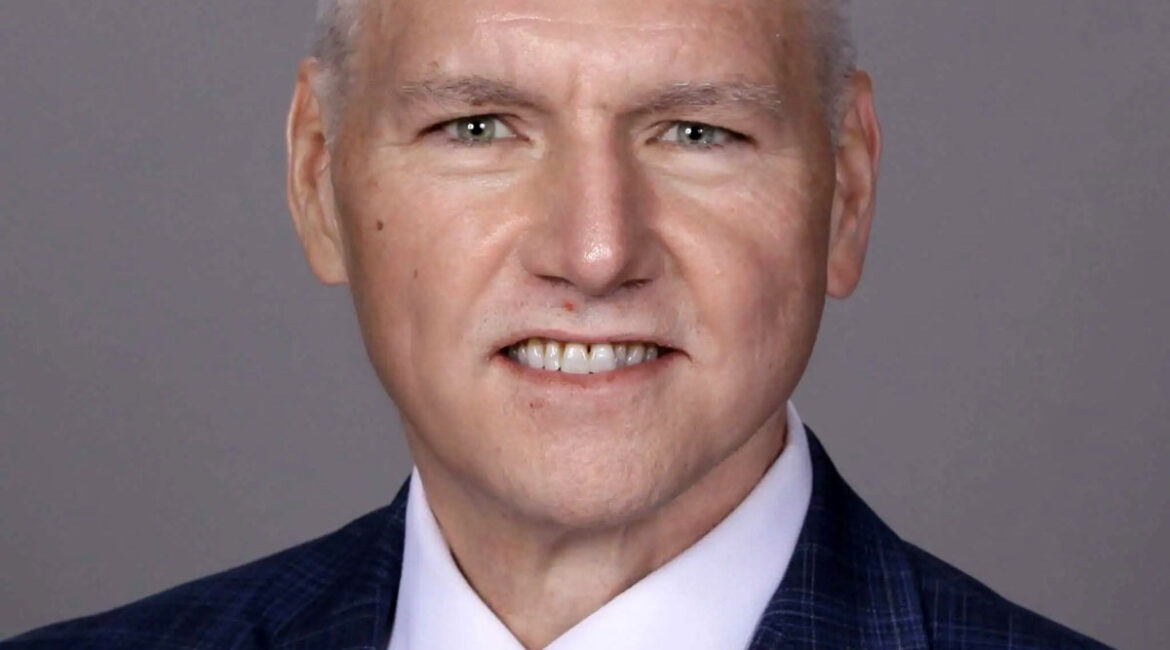 Trauma Solutions: A middle-aged man with short gray hair in a dark plaid suit, white shirt, and pink-blue striped tie poses in front of a plain gray background.