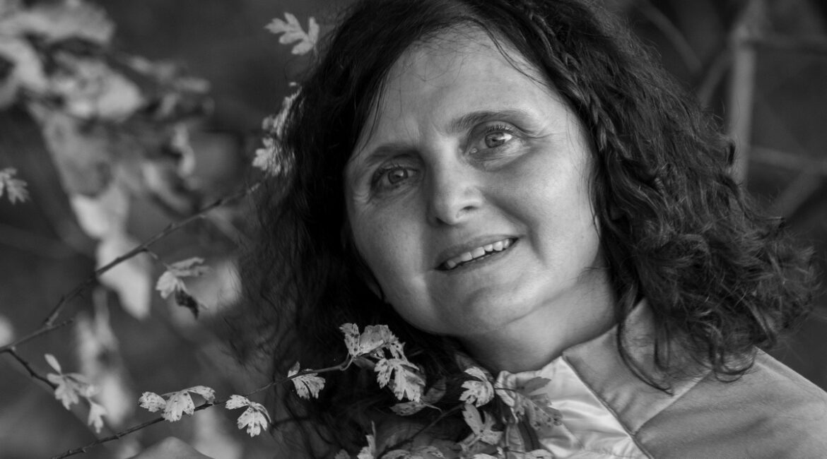 Trauma Solutions: A woman with curly hair and a jacket stands outdoors, holding a leafy branch, with a background of blurred foliage. The image is in black and white.