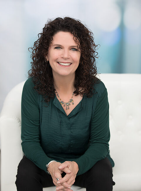 Trauma Solutions: A woman with curly dark hair, wearing a green blouse and necklace, sits and smiles on a white chair with a blurred background.