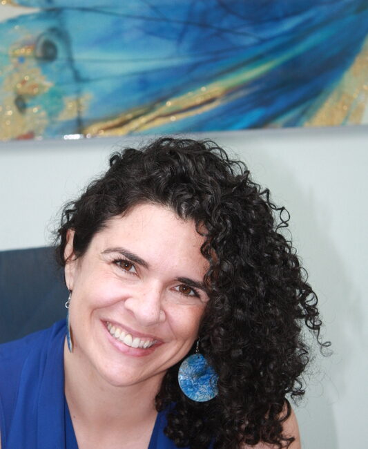 Trauma Solutions: Woman with curly dark hair, wearing a blue top and blue earring, smiling at the camera with an abstract blue artwork in the background.