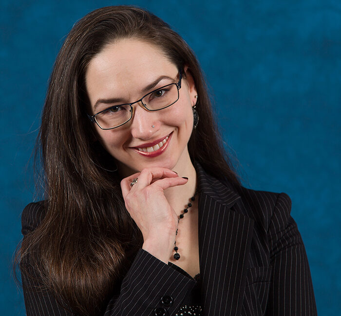 Trauma Solutions: Woman with long brown hair and glasses, wearing a black pinstripe blazer and necklace, smiling with her hand resting on her chin against a blue background.