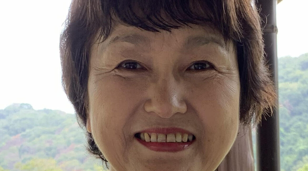 Trauma Solutions: A woman with short dark hair smiles at the camera while standing outdoors, with trees and hills visible in the background.
