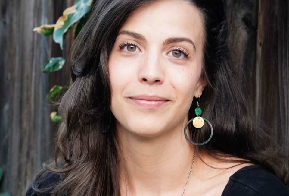 Trauma Solutions: A woman with long brown hair and hoop earrings is sitting outdoors in front of a wooden fence and flowering plants, wearing a black top and looking at the camera.
