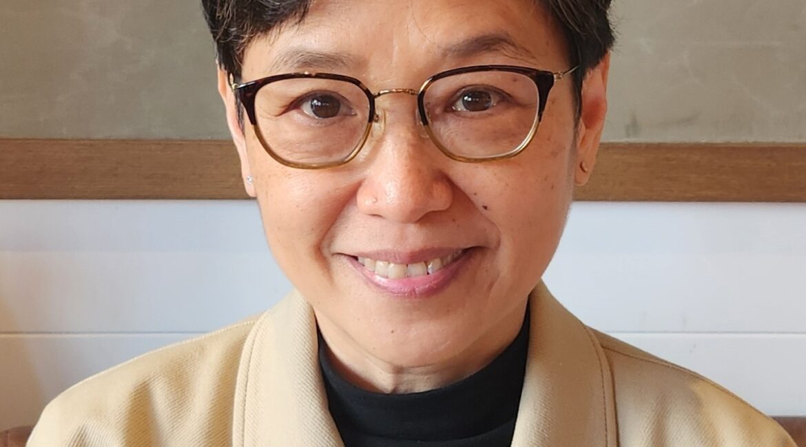 Trauma Solutions: A woman with short dark hair and glasses smiles at the camera, wearing a beige coat over a black top, seated indoors against a neutral background.