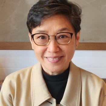 Trauma Solutions: A woman with short dark hair and glasses smiles at the camera, wearing a beige coat over a black top, seated indoors against a neutral background.