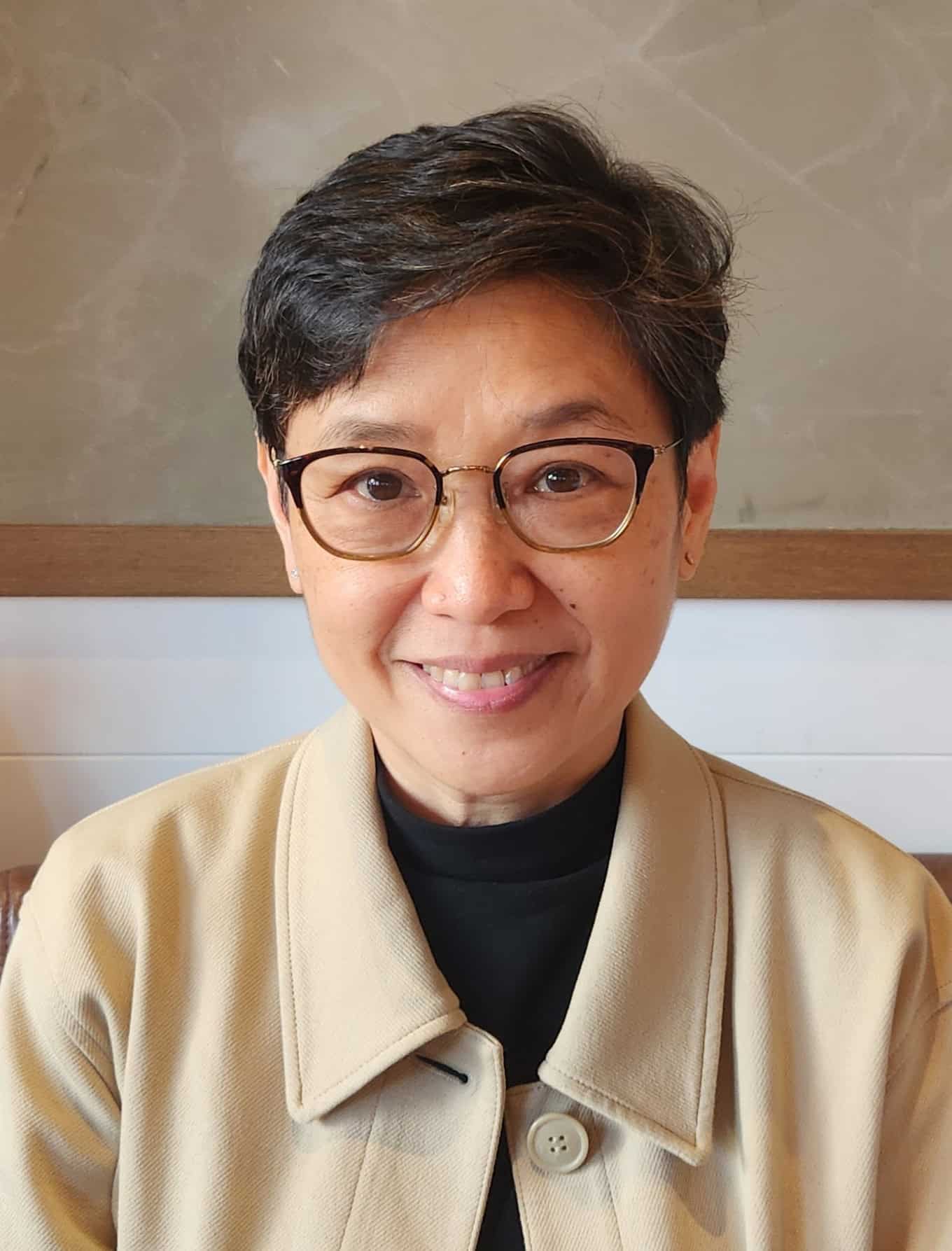 Trauma Solutions: A woman with short dark hair and glasses smiles at the camera, wearing a beige coat over a black top, seated indoors against a neutral background.