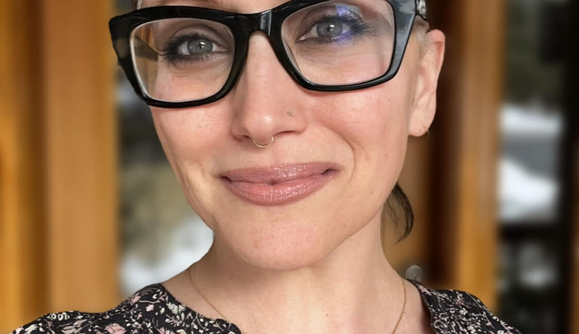 Trauma Solutions: A person with light skin and glasses, wearing a floral top and a necklace, stands in front of a wooden door and smiles at the camera.