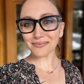 Trauma Solutions: A person with light skin and glasses, wearing a floral top and a necklace, stands in front of a wooden door and smiles at the camera.