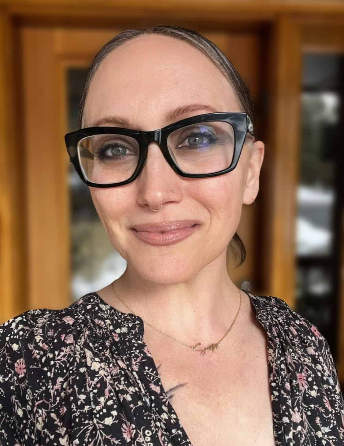 Trauma Solutions: A person with light skin and glasses, wearing a floral top and a necklace, stands in front of a wooden door and smiles at the camera.