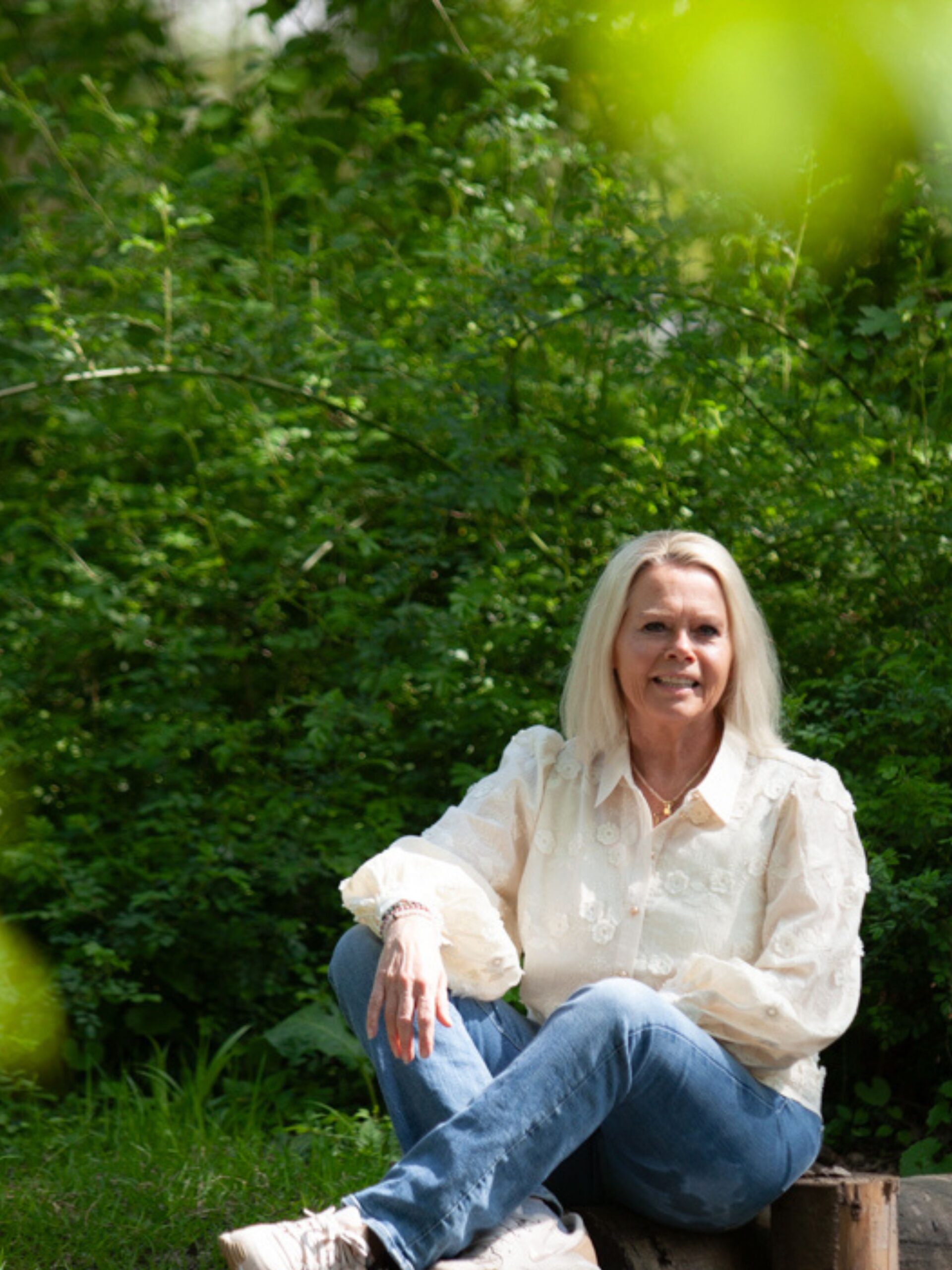 Solutions pour les traumatismes : Une femme aux cheveux blonds mi-longs est assise sur une bûche dans un jardin, vêtue d'un chemisier blanc et d'un jean bleu.