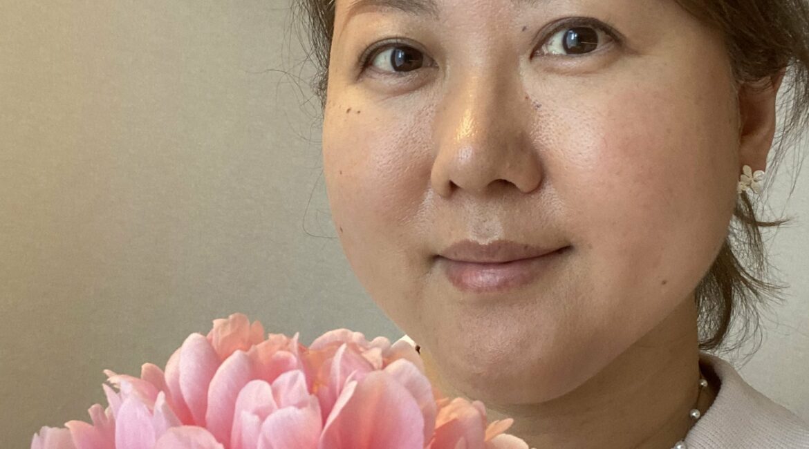 Trauma Solutions: A woman with light brown hair holds a pink peony flower and looks at the camera, against a plain background.