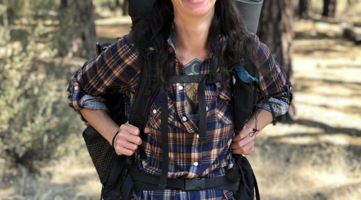 Trauma Solutions: A person with glasses and a red cap, wearing a plaid shirt and shorts, stands smiling in a sunlit forest with a backpack and camping gear.