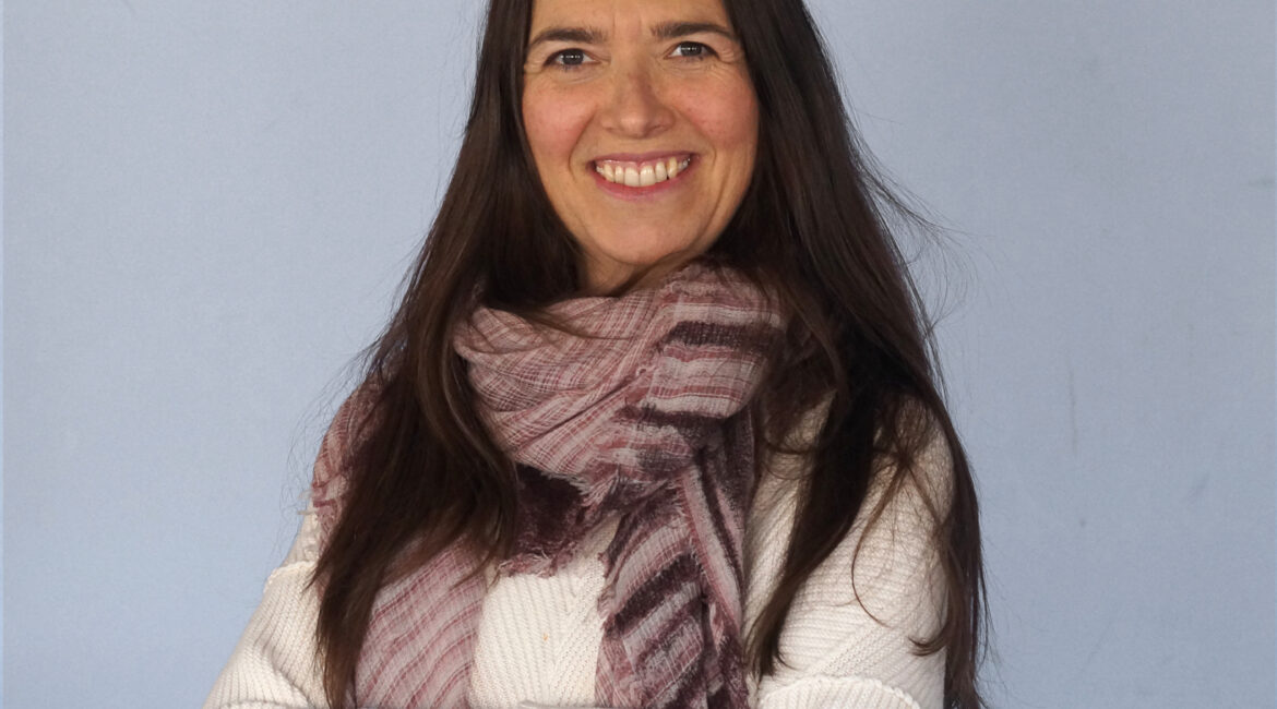 Trauma Solutions: Woman with long dark hair wearing a white sweater and a patterned scarf, standing with arms crossed and smiling in front of a plain light blue background.