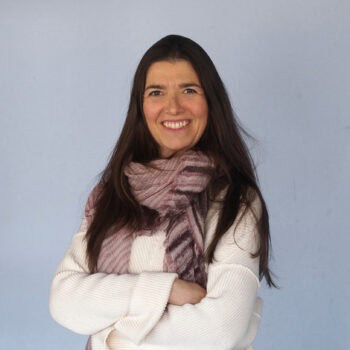 Trauma Solutions: Woman with long dark hair wearing a white sweater and a patterned scarf, standing with arms crossed and smiling in front of a plain light blue background.
