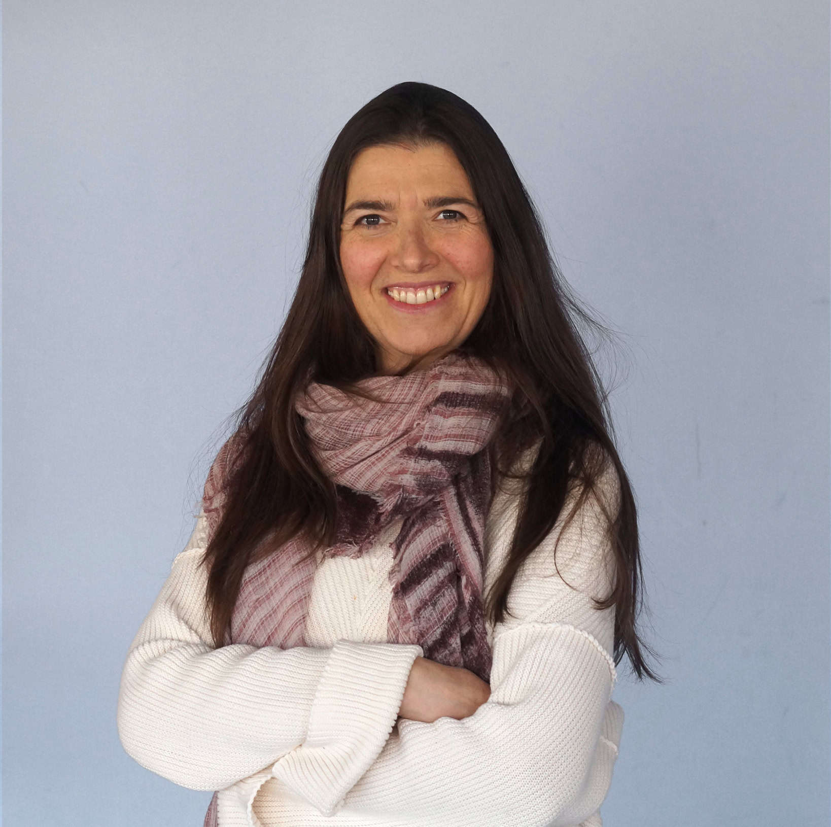 Trauma Solutions: Woman with long dark hair wearing a white sweater and a patterned scarf, standing with arms crossed and smiling in front of a plain light blue background.