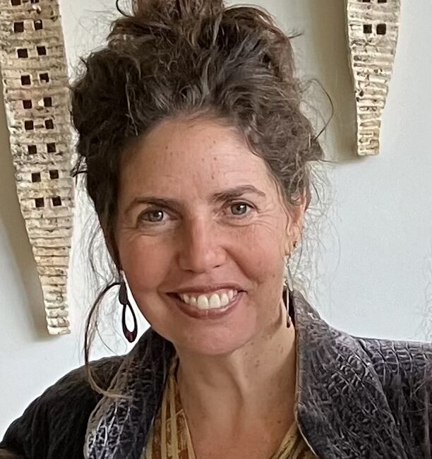 Trauma Solutions: A woman with curly brown hair in an updo and large earrings smiles at the camera, standing in front of a light-colored wall with decorative art pieces.