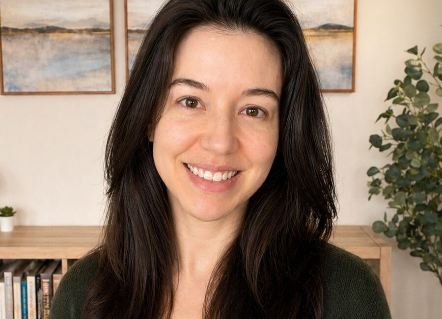 Trauma Solutions: A woman with long dark hair smiles at the camera, standing indoors with bookshelves, framed artwork, and a plant in the background.
