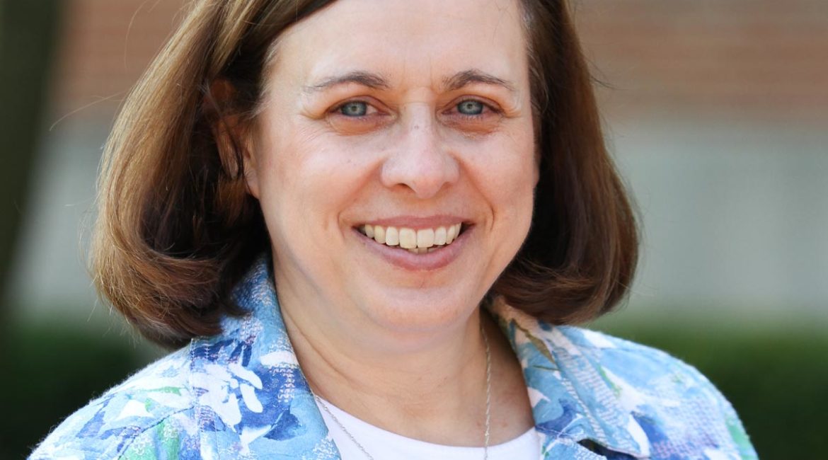 A woman in a floral shirt smiling in front of a brick building.
