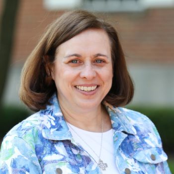 A woman in a floral shirt smiling in front of a brick building.