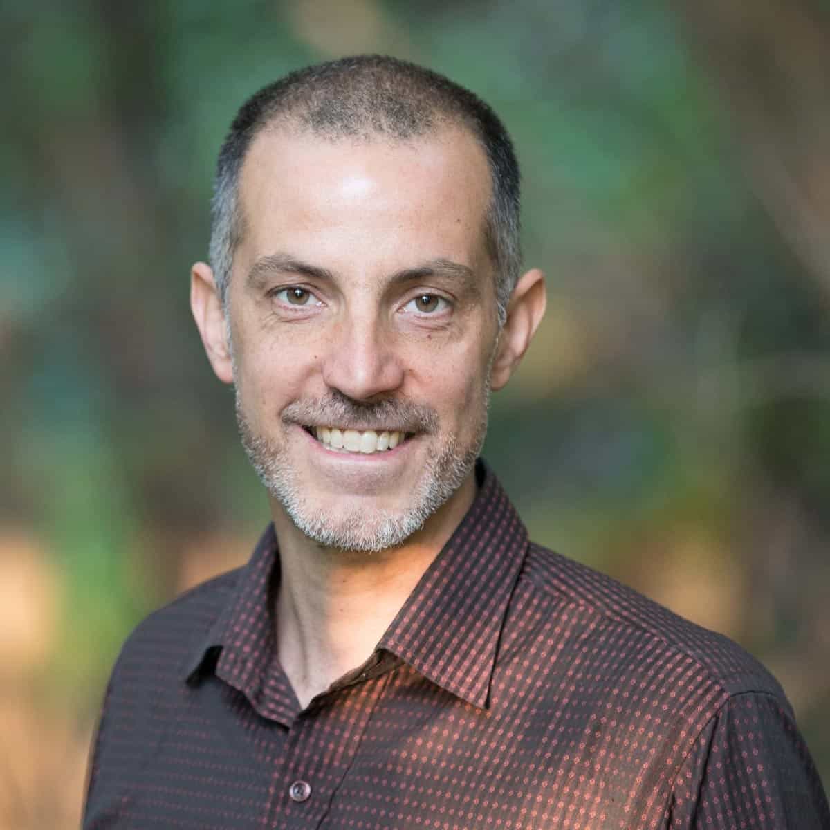 A man in a brown shirt smiling in front of trees.