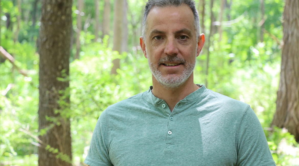 Trauma Solutions: A man with gray hair and a short beard stands outdoors in a green forest, wearing a light green short-sleeve henley shirt.