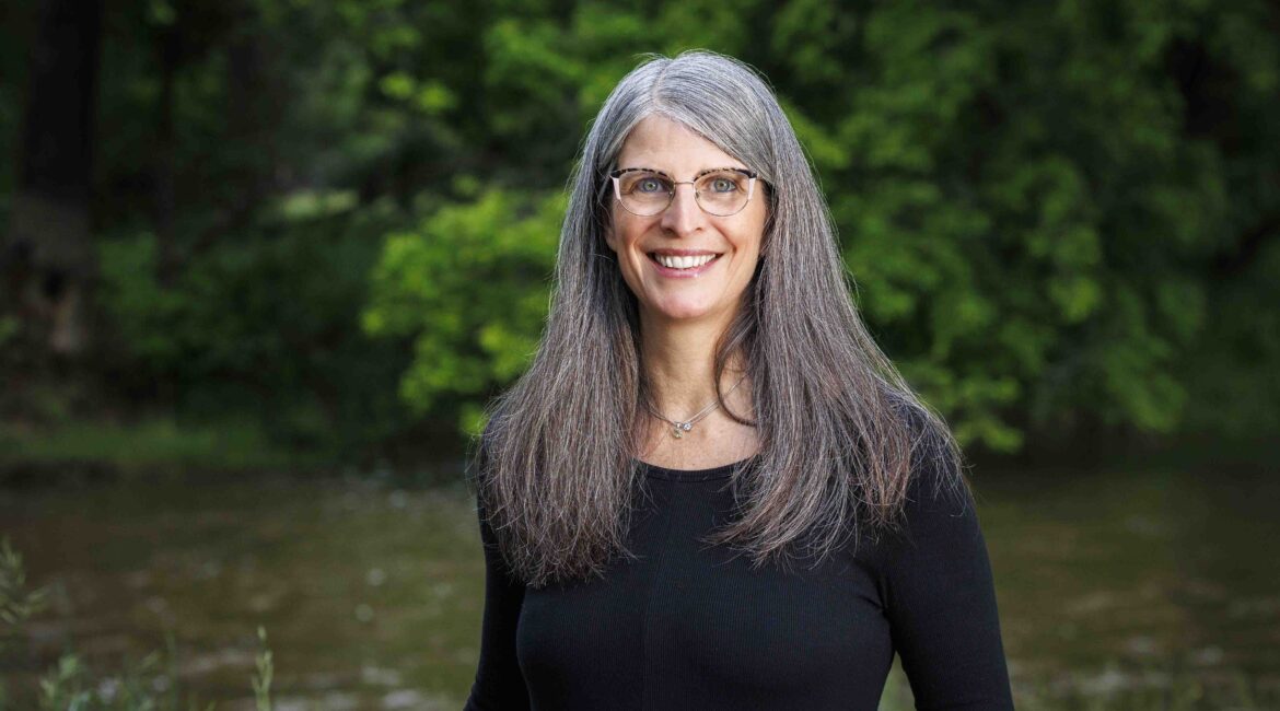 Trauma Solutions: A woman with long gray hair and glasses, wearing a black top, stands outdoors in front of green trees and a river, smiling at the camera.
