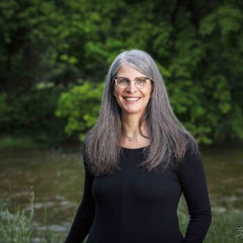 Trauma Solutions: A woman with long gray hair and glasses, wearing a black top, stands outdoors in front of green trees and a river, smiling at the camera.