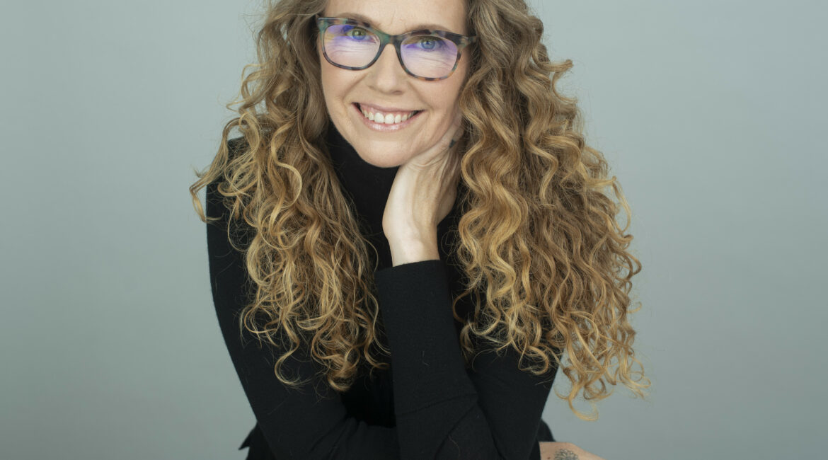 Trauma Solutions: A woman with long curly hair and glasses, wearing a black top, sits at a wooden stool and smiles at the camera with her chin resting on her hand.
