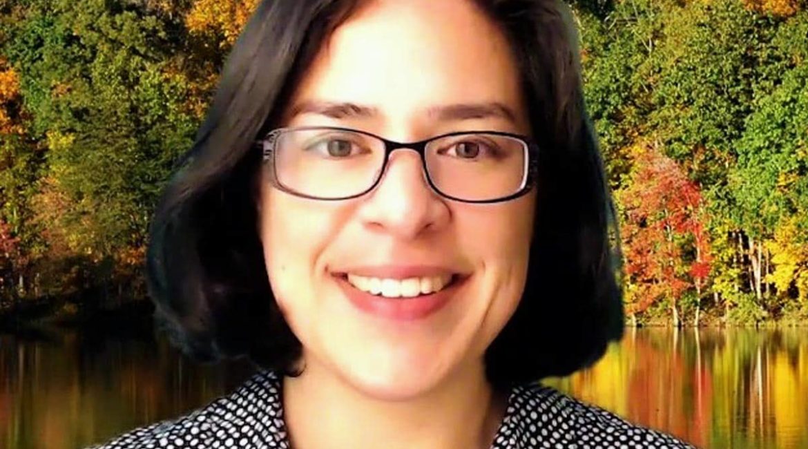 A woman with glasses smiling in front of a lake.