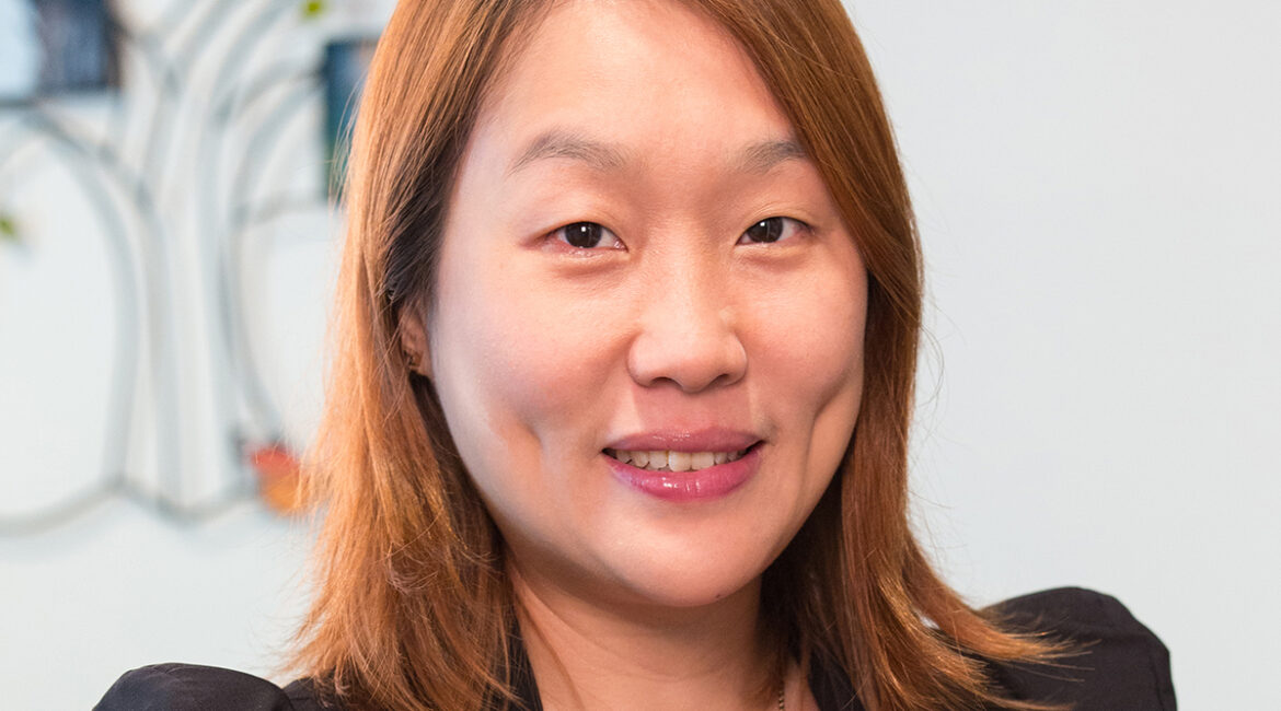 Trauma Solutions: A woman with straight brown hair, wearing a black blazer and red top, smiles at the camera in an indoor setting with a blurred background.