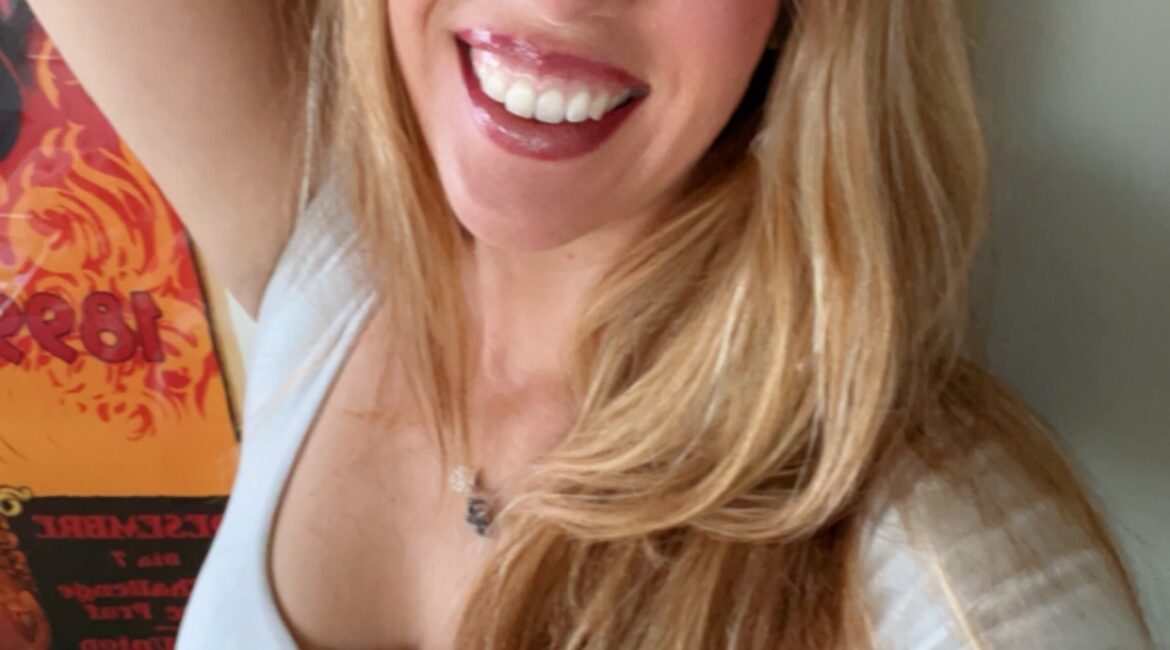 Trauma Solutions: A woman with long blonde hair and a white top smiles at the camera, standing indoors near a wall with a colorful poster in the background.