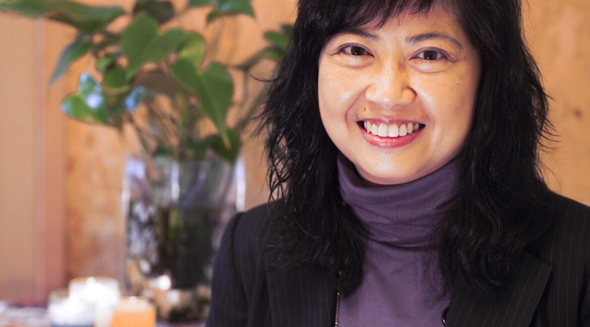 Trauma Solutions: A woman with black hair in a dark blazer and purple turtleneck smiles at the camera; there are plants and lit candles in the background.
