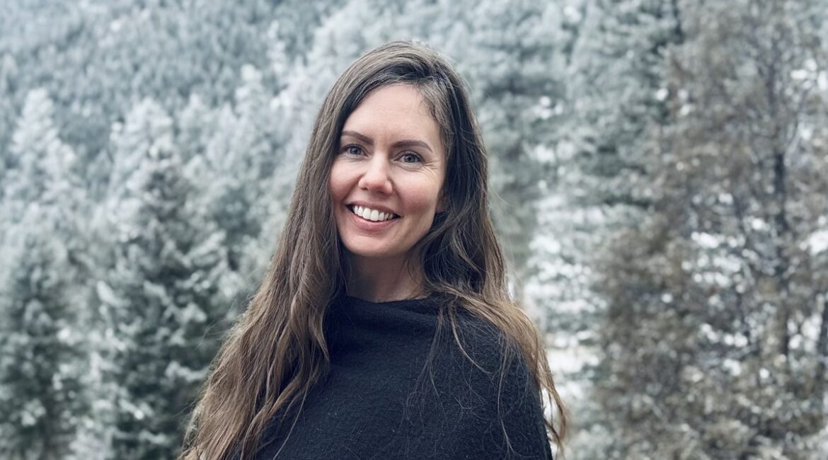 Solutions pour les traumatismes&nbsp;: Femme aux longs cheveux bruns portant un châle sombre, souriant à l’extérieur devant des arbres enneigés et des montagnes boisées.