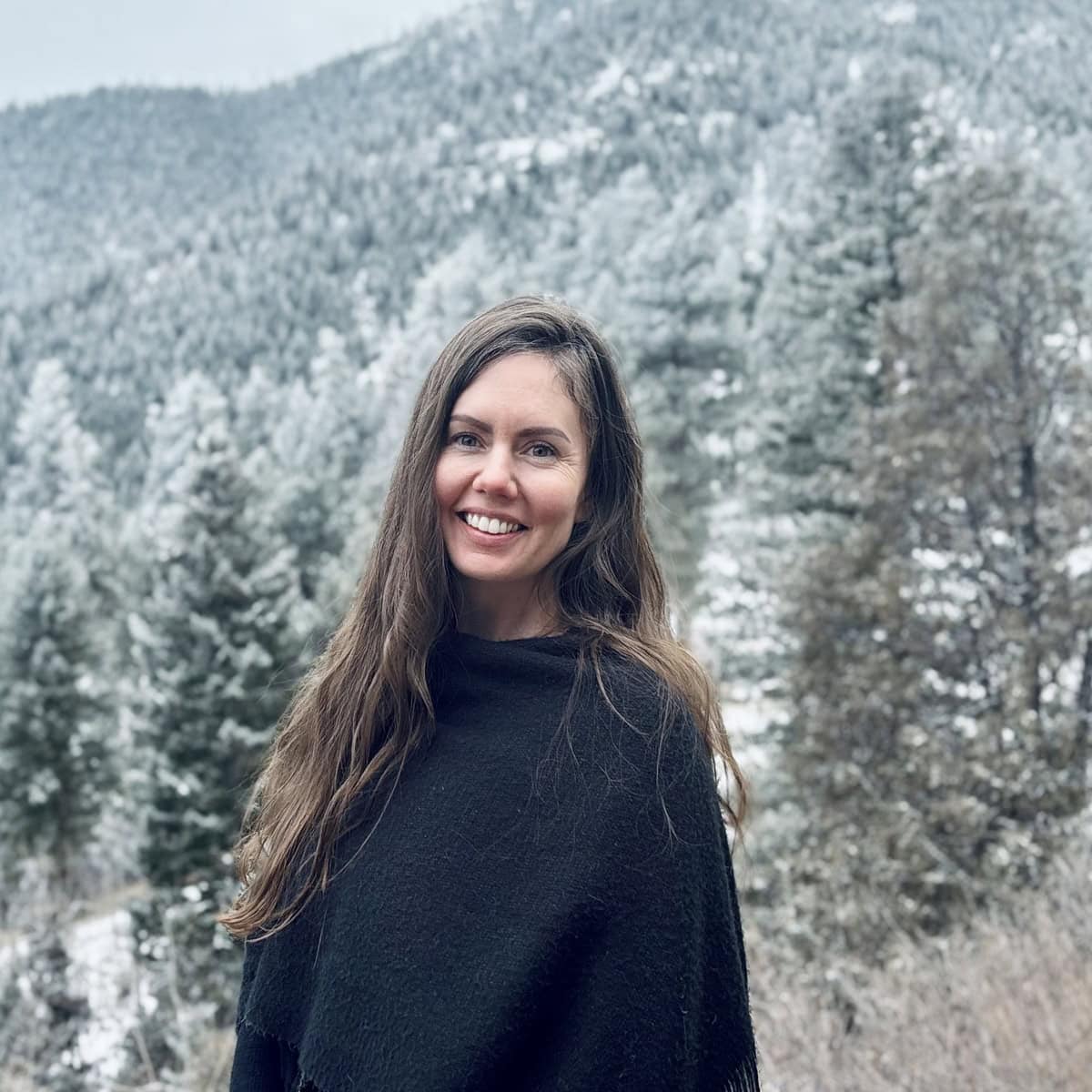 Solutions pour les traumatismes&nbsp;: Femme aux longs cheveux bruns portant un châle sombre, souriant à l’extérieur devant des arbres enneigés et des montagnes boisées.