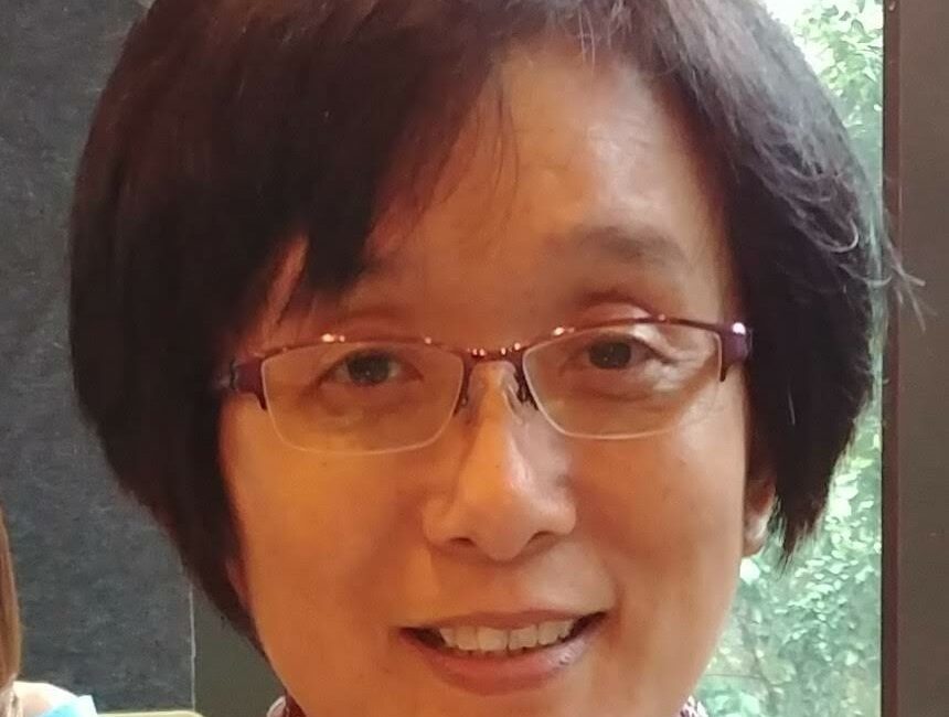 Trauma Solutions: A woman with short dark hair and glasses smiles at the camera; she is wearing a light-patterned scarf, seated indoors near a window.