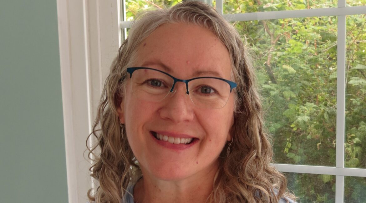 Trauma Solutions: A woman with curly gray hair and glasses smiles in front of a window with greenery outside.