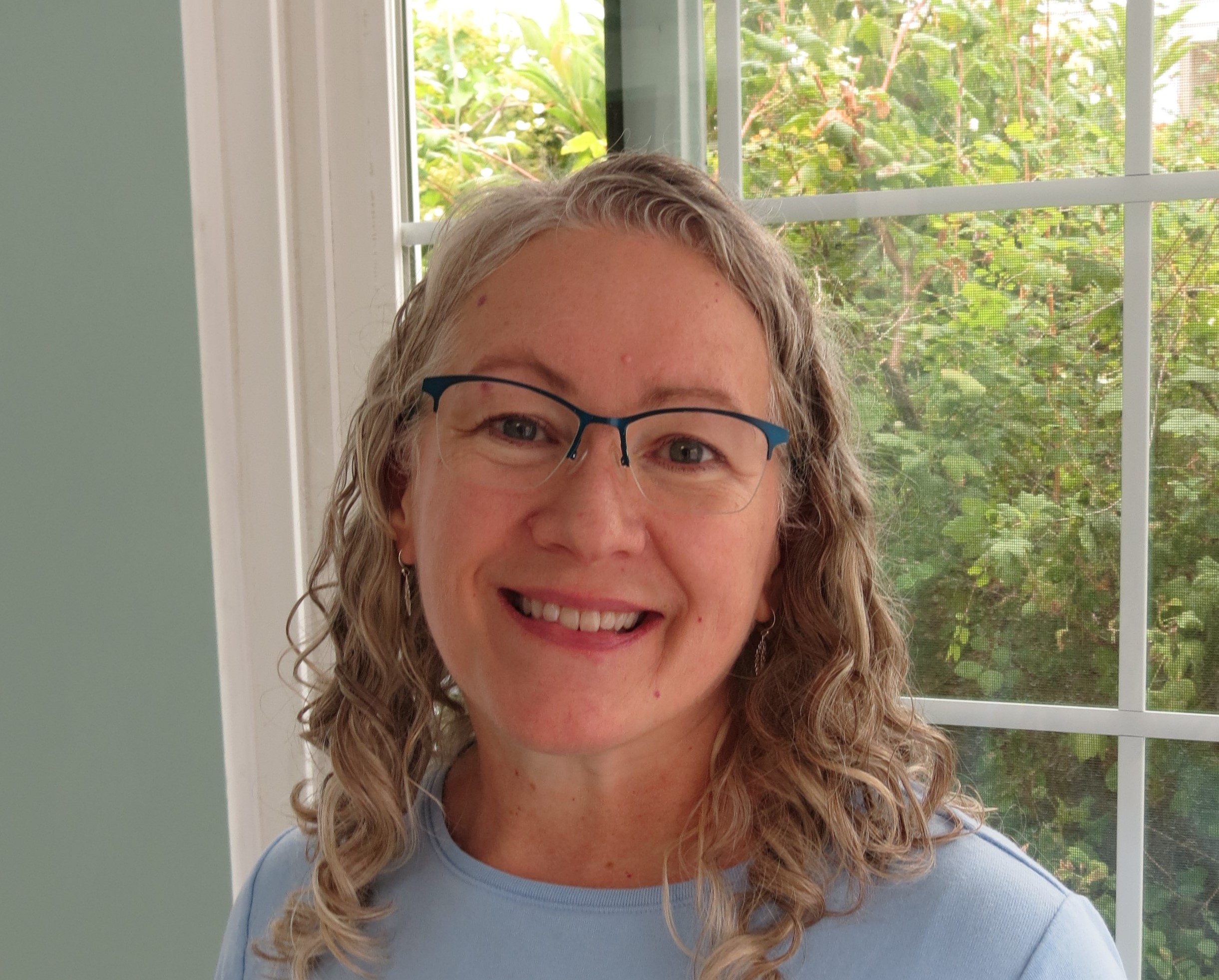 Trauma Solutions: A woman with curly gray hair and glasses smiles in front of a window with greenery outside.