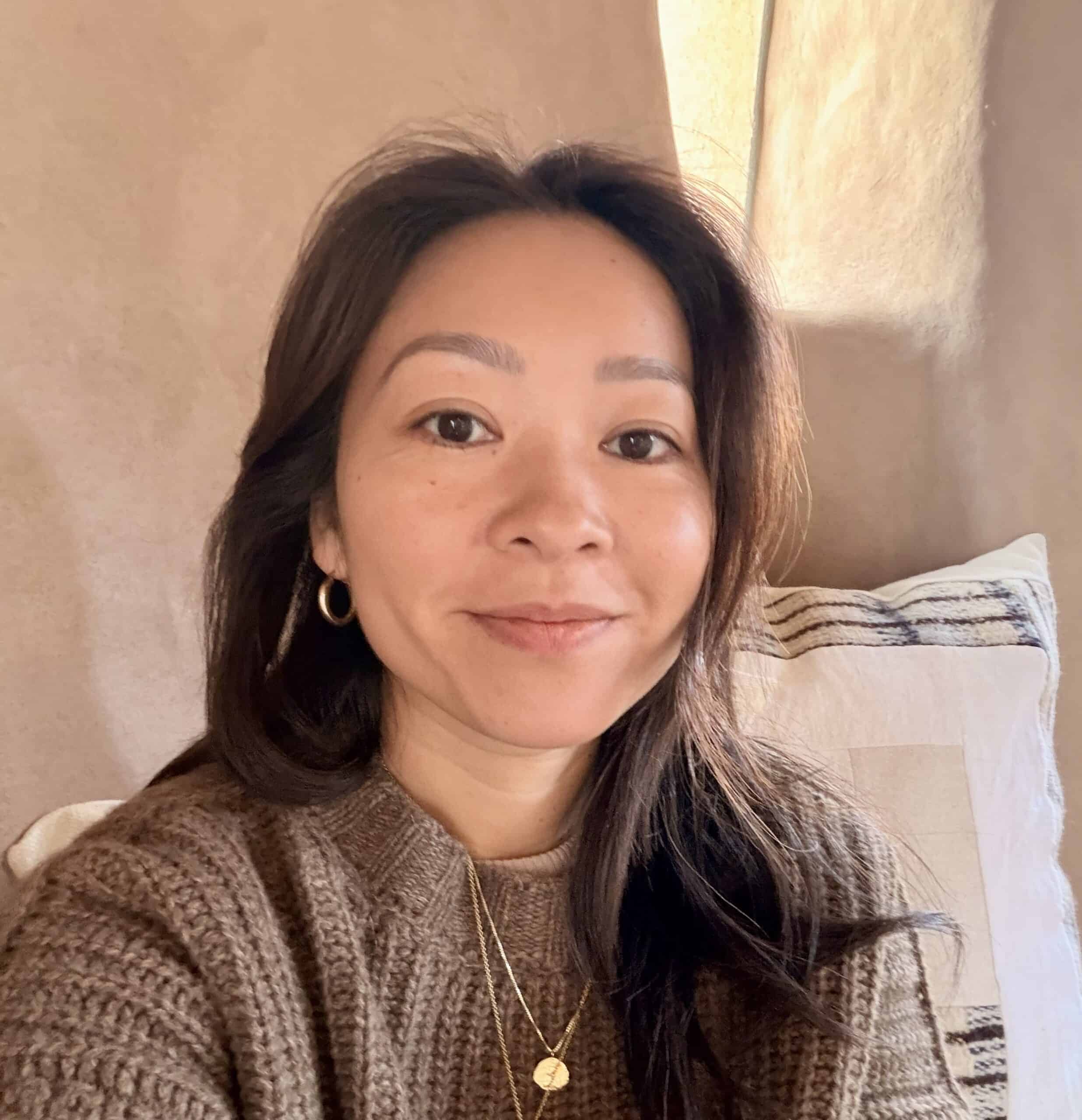 Soluciones para el trauma: Una mujer de cabello largo y oscuro, vestida con un suéter marrón y joyas de oro, está sentada en el interior de una vivienda, frente a una pared beige y un cojín a rayas, mirando a la cámara con una leve sonrisa.